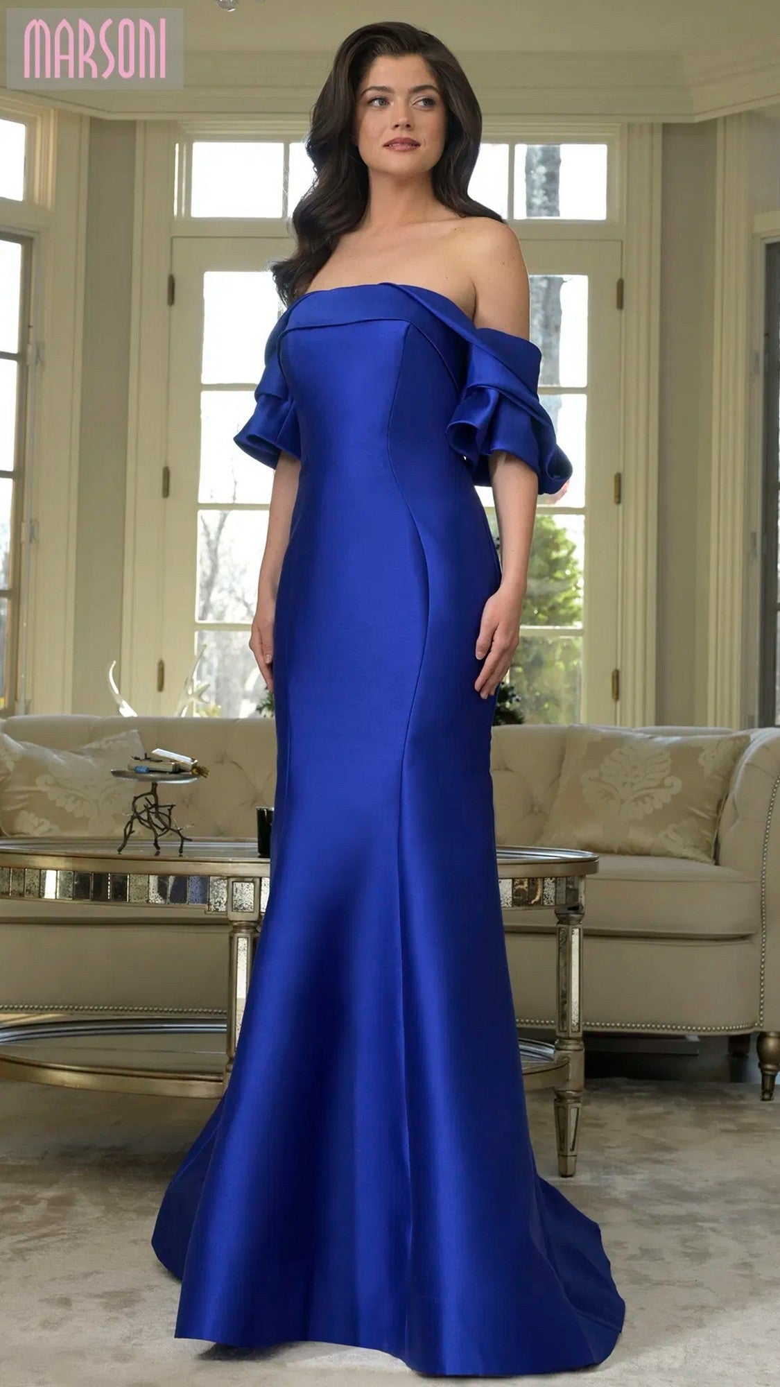 A woman models the Marsoni MV1290 long formal dress in royal blue, featuring a mermaid silhouette, as she stands in a living room with large windows and beige furniture.